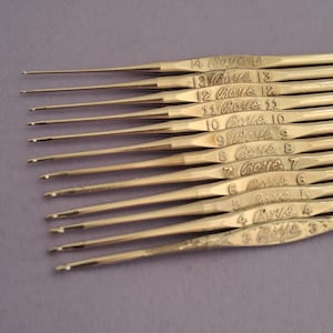 Op de afbeelding: Close-up van een set vintage haaknaalden. De haken zijn gemaakt van een goudkleurig metaal en zijn in parallelle lijnen gerangschikt. Elke haak is gemarkeerd met een nummer en het woord "Boys".