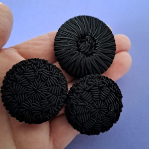 Könnte beinhalten: Drei schwarze, runde Knöpfe mit detaillierten, strukturierten Mustern. Die Knöpfe bestehen aus einem dicht gewebten Material, das ein detailliertes Oberflächendesign erzeugt. Die Knöpfe variieren leicht in Größe und Design und bieten ein einzigartiges Set für Näh- oder Bastelprojekte.