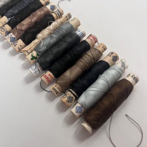 May include: A selection of thread spools in a range of neutral colours, including grey, brown, and black. Each spool features a paper label with text and a decorative design. The threads are neatly wound, suitable for sewing and craft activities.