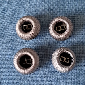 Puede incluir: Cuatro botones cubiertos de tela plateada con un centro negro y dos bucles de metal plateado.