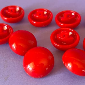 Pode incluir: Uma coleção de botões vermelhos redondos e brilhantes. Cada botão apresenta uma superfície superior lisa e abobadada e uma haste elevada na parte inferior com um pequeno orifício para fixação. Os botões estão dispostos de forma dispersa.