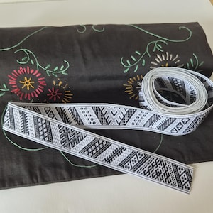 Könnte beinhalten: Eine Rolle weißes Webband mit einem sich wiederholenden schwarzen geometrischen Muster aus diagonalen Linien und Dreiecken. Das Band liegt auf einem dunkelbraunen Stoffhintergrund, der mit gestickten Blumenmotiven in Rot, Gelb und Hellgrün verziert ist.