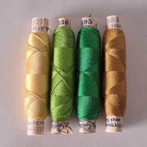 Pode incluir: Quatro carretéis de linha em tons de ouro e verde. Os carretéis têm etiquetas de papel com números e texto. As cores da linha incluem ouro claro, verde limão, verde floresta e um ouro mais escuro. Os rótulos mostram a metragem e a marca da linha.