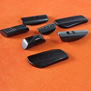 Op de afbeelding: Een set van zeven zwarte knopen, sommige met twee gaten en sommige met één gat. De knopen zijn gemaakt van een glad, glanzend materiaal en hebben verschillende vormen, waaronder rechthoekig, ovaal en afgerond.