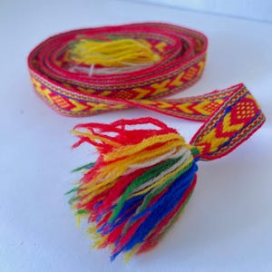 May include: A woven fabric belt featuring a repeating geometric pattern in red, yellow, and blue. The belt is coiled and finished with a multi-colored tassel at the end, consisting of red, yellow, white, green, and blue yarn strands.