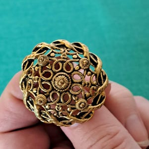 May include: A gold-toned ring with a filigree design. The ring features a circular center with a floral pattern and is surrounded by a twisted rope design.