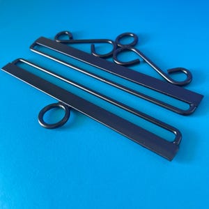 May include: Two matte black metal wall hanging brackets. Each bracket features a long, flat horizontal bar with a rectangular slot, complemented by decorative scrollwork or a circular ring attachment for mounting or displaying items securely.