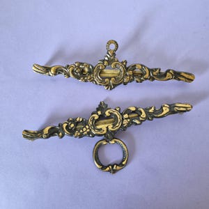 Peut inclure: Deux poignées de meuble en métal doré, ornées de motifs floraux en relief. La poignée supérieure présente un petit anneau de fixation central, tandis que la poignée inférieure est dotée d'un anneau de tirage circulaire plus grand.