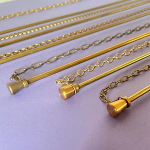 May include: Various gold-coloured chains and rods, some with decorative features and small cone-shaped finials. The chains have different styles, including link and beaded designs. The items are displayed on a light purple surface.