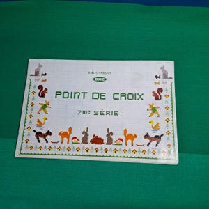 Pode incluir: Um livreto branco com um padrão de ponto cruz colorido na capa. O título é "Point de Croix" com o logotipo da DMC e "7ime Serie" abaixo. O padrão apresenta animais como esquilos, coelhos e pássaros.