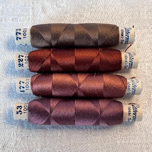 Op de afbeelding: Vier spoelen Gütermann Ideal-Naai draad in de kleuren bruin, rood, bordeaux en donker bordeaux. De spoelen zijn gelabeld met de nummers 773, 227, 477 en 53, en de tekst "Gütermann Ideal-Naai 100/3".