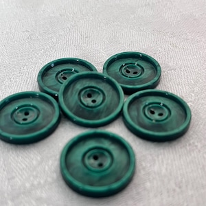 Puede incluir: Seis botones de plástico verde con dos agujeros cada uno. Los botones tienen un efecto marmoleado y están dispuestos en semicírculo.