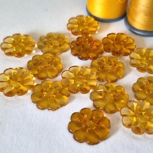 Peut inclure: Une collection de boutons en plastique translucide de couleur ambre en forme de fleurs à huit pétales arrondis. Les boutons sont dispersés sur une surface blanche, avec deux bobines de fil jaune visibles dans le coin supérieur droit de l'image.