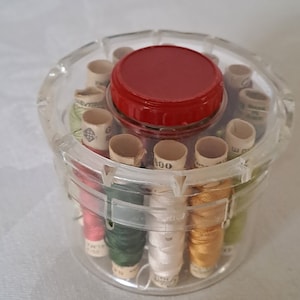 May include: A clear plastic container with a red lid holds spools of thread. The spools are arranged in a circle around the container. The thread is in various colours, including red, green, white, and yellow.