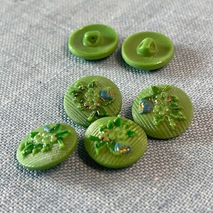 Könnte beinhalten: Eine Auswahl an grünen Knöpfen in verschiedenen Größen und Designs. Einige sind rund und schlicht, andere weisen ein geprägtes Blumenmuster mit blauen und roten Akzenten auf. Die Knöpfe liegen auf einer strukturierten, hellblauen Oberfläche.