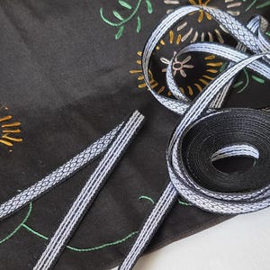 Könnte beinhalten: Eine schwarze Stoffrolle mit einem gewebten weißen geometrischen Muster an den Rändern, präsentiert zusammen mit einem Stück des gleichen Bandes. Das Band liegt auf einem schwarzen Textiluntergrund mit bunter Blumenstickerei in Grün, Rot und Gelb.