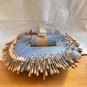 Pode incluir: Uma almofada azul, oval, para fazer renda, com inúmeras bobinas de madeira e fio branco. A almofada tem uma estrutura central de madeira e uma borda de renda decorativa. As bobinas estão dispostas ao redor da borda, prontas para criar renda.