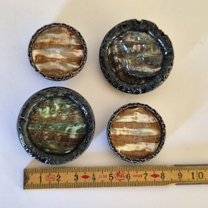 Puede incluir: Cuatro botones circulares con centros de concha de abulón iridiscente y bordes de metal oscuro texturizado. Una regla de madera amarilla está colocada en la parte inferior, mostrando que el botón más grande mide 3,8 cm y el más pequeño 2,5 cm.