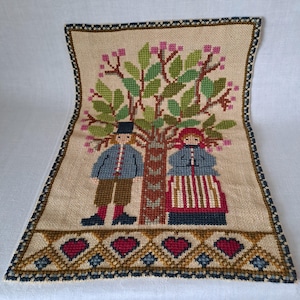 May include: A vintage needlepoint wall hanging featuring a stylized tree with two figures in blue and red clothing. The figures are standing in front of the tree, which has a brown trunk and green leaves. The background is a beige fabric with a brown border.
