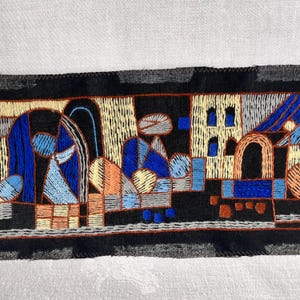 May include: Embroidered textile with a geometric design in blue, orange, yellow, and gray on a black background. The design features abstract shapes and architectural elements, stitched with various thread colors. The textile is set against a white fabric.
