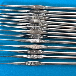 Op de afbeelding: Een verzameling van twaalf zilverkleurige metalen haaknaalden die horizontaal zijn gerangschikt. Elke naald heeft een afgeplat middengedeelte met de tekst U.S.A. en verschillende numerieke waarden zoals 10¢, 15¢, 25¢ en 35¢.