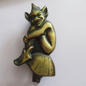 Puede incluir: Una figurita de metal de color dorado de un duende o diablillo sonriente sentado sobre una base. La figurita está detallada con un sombrero puntiagudo, orejas y una expresión traviesa.