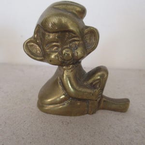 May include: A brass gnome figurine in a seated position, wearing a pointed hat and with large ears. The gnome is relaxed, with crossed arms and one leg extended. The figurine has a polished finish.