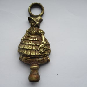 Könnte beinhalten: Ein Türklopfer aus Messing in Form eines Mädchens, das ein Kleid und einen Hut trägt. Der Türklopfer hat oben einen Ring und der Name "DOREEN" ist unten eingraviert.