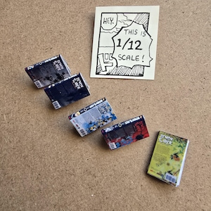 May include: Miniature "Demon Slayer" manga books in various colors, arranged on a brown surface. The books are labeled and detailed, with a handwritten sign stating "This is 1/12 scale!" in a comic-style frame.