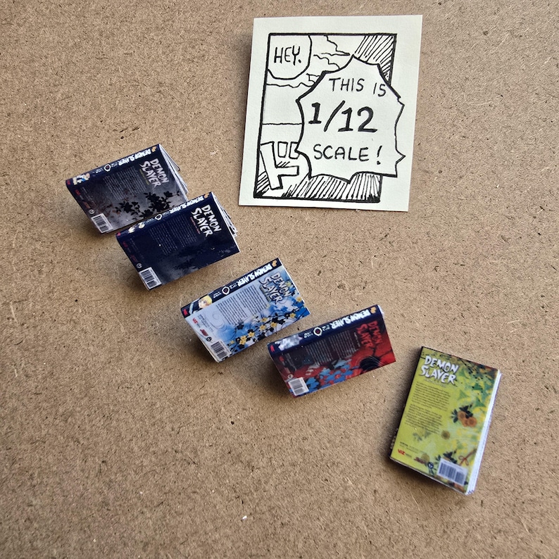 May include: Miniature "Demon Slayer" manga books in various colors, arranged on a brown surface. The books are labeled and detailed, with a handwritten sign stating "This is 1/12 scale!" in a comic-style frame.