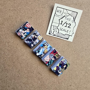 May include: A collection of miniature "Demon Slayer" manga books with colorful covers, arranged diagonally on a brown surface. A hand-drawn sign reads "HEY. THIS IS 1/12 SCALE!" in a speech bubble.