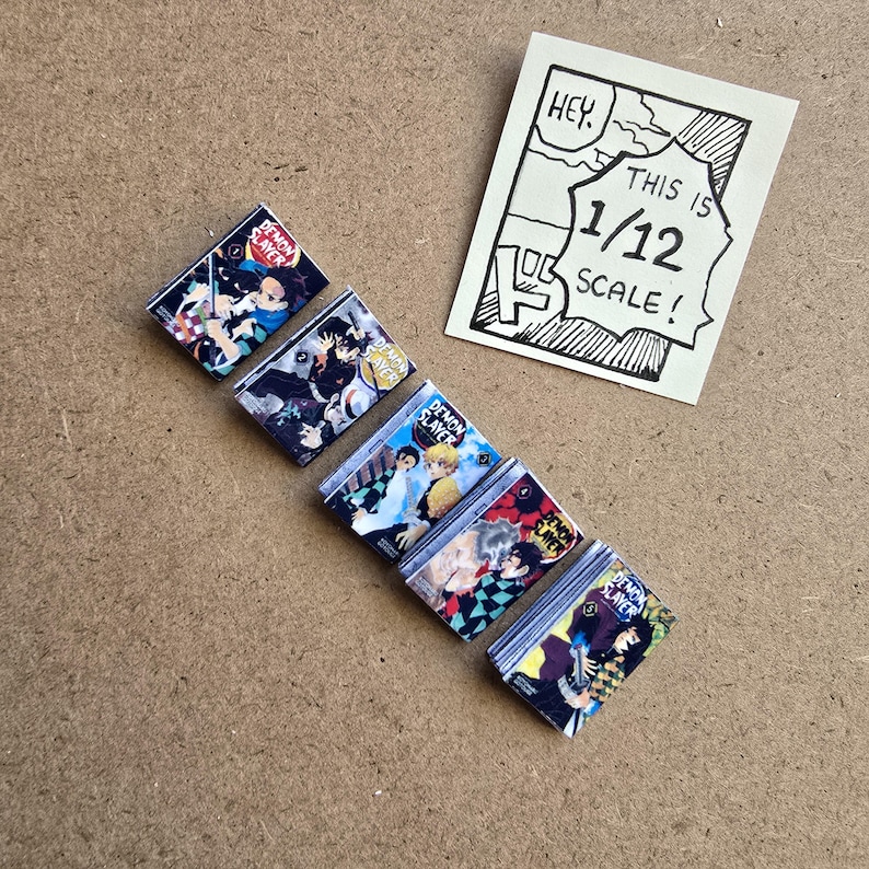 May include: A collection of miniature "Demon Slayer" manga books with colorful covers, arranged diagonally on a brown surface. A hand-drawn sign reads "HEY. THIS IS 1/12 SCALE!" in a speech bubble.