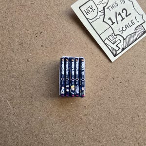 May include: A stack of miniature manga books with the title "Demon Slayer" visible on the spines. The books are dark blue with colorful illustrations. A small, hand-drawn sign reads "Hey. This is 1/12 scale!"