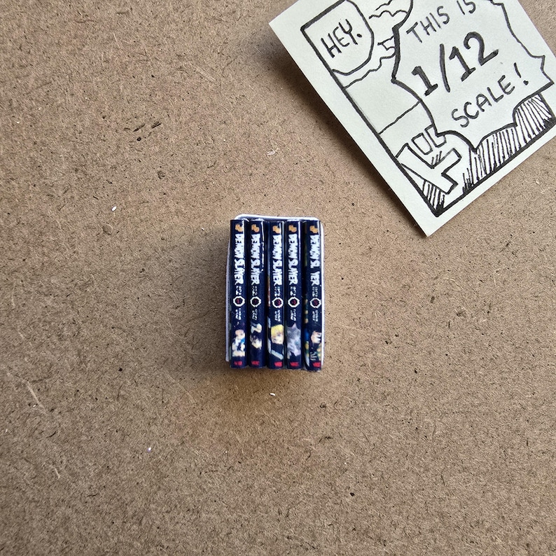 May include: A stack of miniature manga books with the title "Demon Slayer" visible on the spines. The books are dark blue with colorful illustrations. A small, hand-drawn sign reads "Hey. This is 1/12 scale!"
