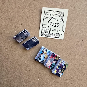 May include: Miniature comic books with colorful covers, featuring characters and the text "DEMON SLAYER." Two stacks of books are visible, along with two individual books and a small sign that reads "HEY. THIS IS 1/12 SCALE!"