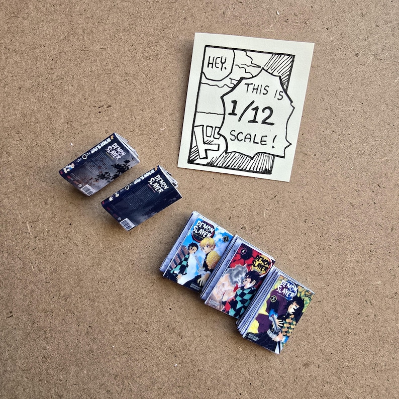 May include: Miniature comic books with colorful covers, featuring characters and the text "DEMON SLAYER." Two stacks of books are visible, along with two individual books and a small sign that reads "HEY. THIS IS 1/12 SCALE!"