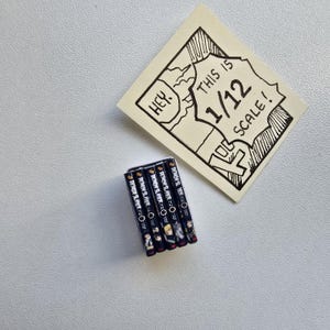 May include: A stack of miniature manga books with the title "Demon Slayer" visible. A handwritten note says "Hey. This is 1/12 scale!" in a comic book style. The books are black with white text and colorful illustrations.