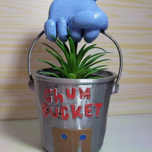 Puede incluir: Un macetero de cubo gris impreso en 3D con una mano azul sosteniendo una planta verde. El cubo tiene las palabras "GHUM BUCKET" impresas en el lateral en letras rojas.