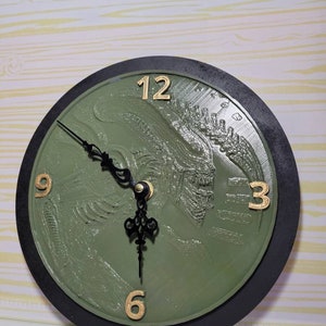 May include: A green clock with a black outer ring and gold numbers. The clock face features a 3D embossed image of an alien creature.