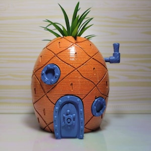 May include: A 3D printed pineapple-shaped house with a blue door and porthole windows. The house is decorated with a green plant on top.