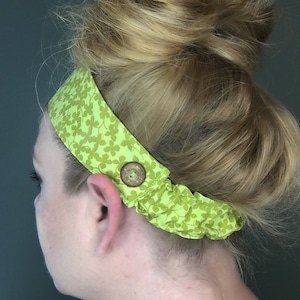 May include: A light green headband with a clover pattern and a wooden button closure. The headband is made of fabric and is worn around the head. The hair is styled in a bun.