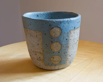 May include: A handmade ceramic cup with a speckled gray body and a light blue rim. The cup features a rectangular blue design with three circular accents. The cup is sitting on a wooden surface.
