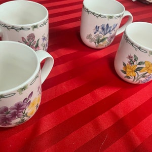 May include: Four white ceramic mugs with floral designs. The mugs have green handles and are decorated with different floral patterns in shades of pink, yellow, blue, and purple.