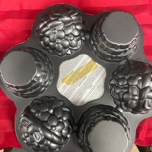May include: A dark gray, non-stick muffin pan with six uniquely shaped molds. Some molds feature floral designs, while others have a ribbed texture. The pan sits on a red surface.
