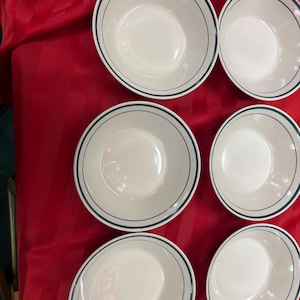 May include: Six white bowls with a blue trim are arranged on a red surface. The bowls are round and have a simple, classic design. The blue trim adds a subtle pop of color to the otherwise plain white bowls. These bowls are suitable for serving soups, salads, or other dishes.