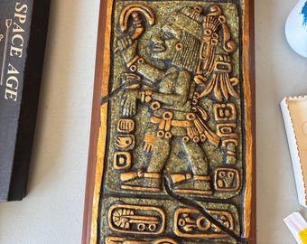 May include: A rectangular, carved relief sculpture with a Mayan figure. The figure is depicted in a detailed, golden-brown relief against a textured, green background. The sculpture is framed in a dark brown wooden border. The sculpture is cracked.