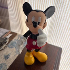 Puede incluir: Una figura de Mickey Mouse en una pose cl&aacute;sica. La figura tiene orejas negras, un cuerpo negro, pantalones cortos rojos con botones blancos, zapatos amarillos y guantes blancos. La figura est&aacute; sobre una superficie marr&oacute;n oscuro.