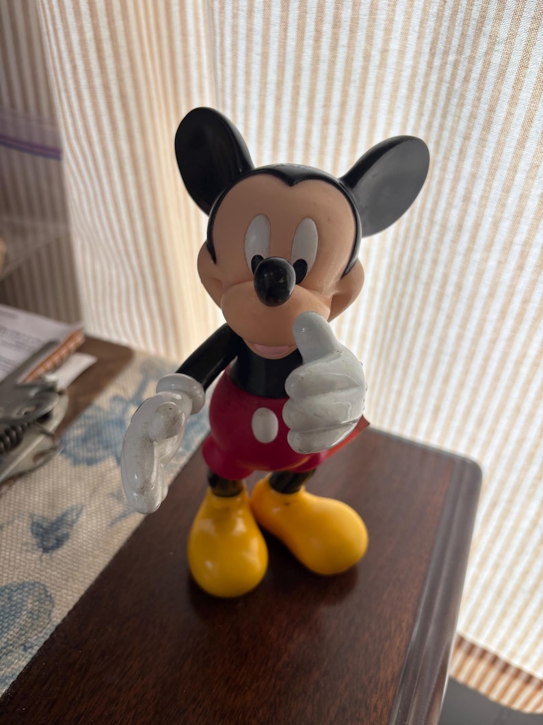 Puede incluir: Una figura de Mickey Mouse en una pose cl&aacute;sica. La figura tiene orejas negras, un cuerpo negro, pantalones cortos rojos con botones blancos, zapatos amarillos y guantes blancos. La figura est&aacute; sobre una superficie marr&oacute;n oscuro.