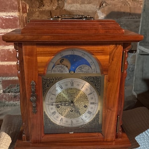 May include: A wooden mantel clock with a rectangular shape. The clock features a gold-colored face with Roman numerals and ornate detailing. The clock's body is made of reddish-brown wood, and it has a handle on top.