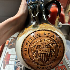 May include: A ceramic jug with a brown and white glaze. The jug has a wooden lid with a metal latch. The jug is decorated with a wooden carving of the United States Navy seal. The seal is surrounded by a rope design and the words "United States Navy".